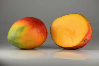 Kokonainen punavihreä mango ja leikattu mango, jonka oranssi hedelmäliha näkyy, harmaalla taustalla