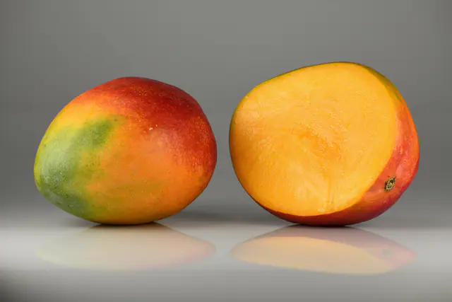 Kokonainen punavihreä mango ja leikattu mango, jonka oranssi hedelmäliha näkyy, harmaalla taustalla