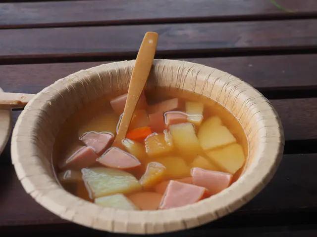 Nakkikeittoa perunoiden, porkkanan ja nakkipalojen kera kartonkikulhossa tummalla puupöydällä