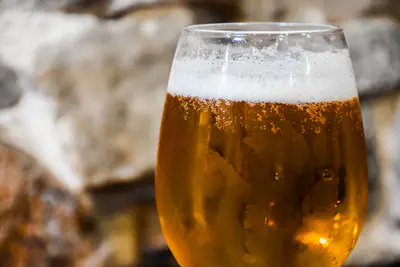 Kylmä vaalea olut lasissa, pinnalla vaahtoa ja kuplia, taustalla sumea kiviseinä