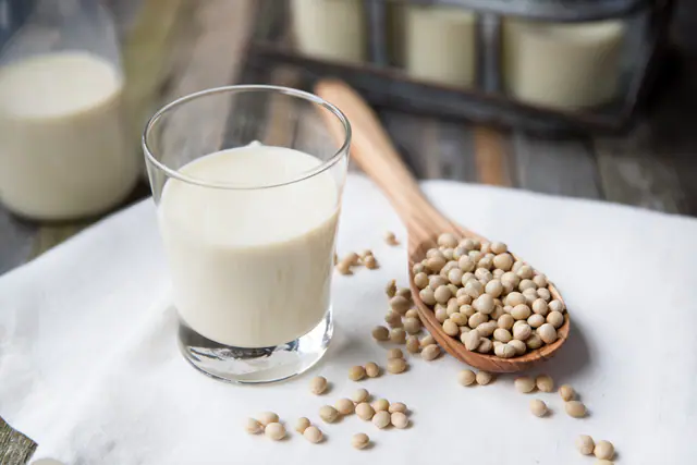 Lasi soijajuomaa valkoisella liinalla, vieressä puulusikallinen kuivattuja soijapapuja
