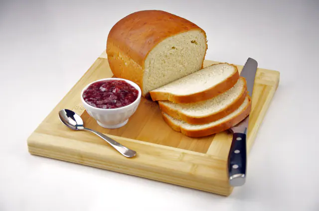Vaalea vehnäleipä viipaloituna leikkuulaudalla, vieressä kulhollinen mansikkahilloa