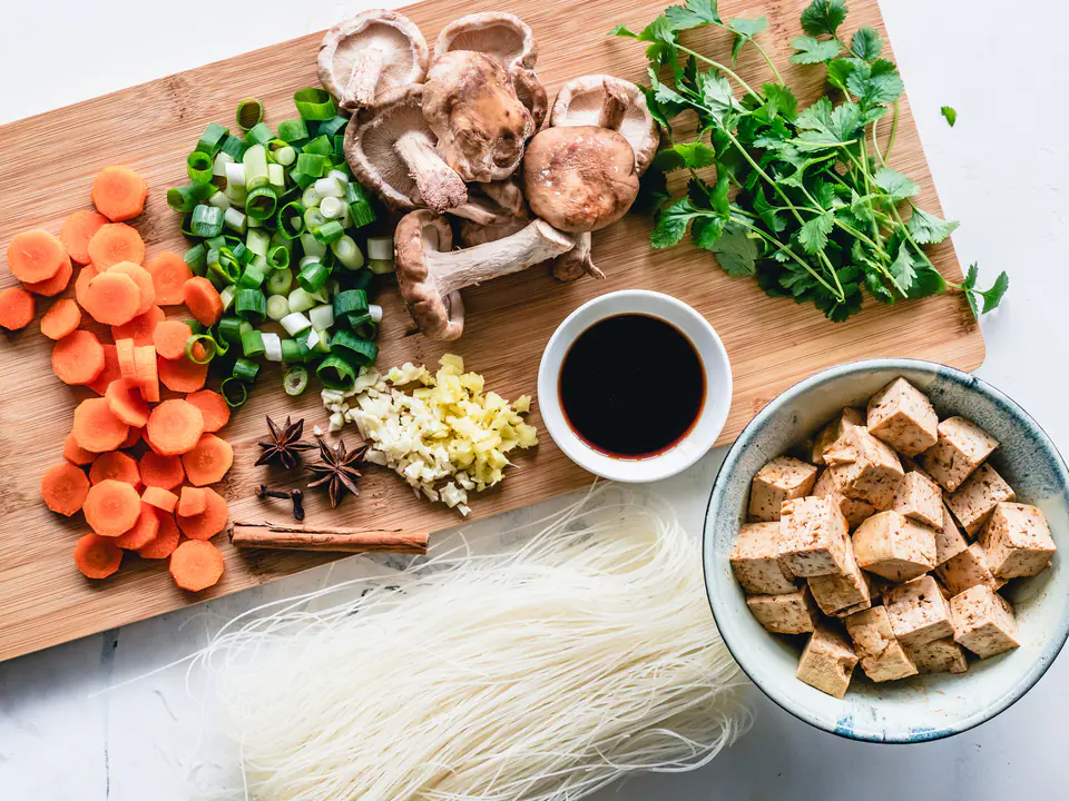 Leikkuulaudalla porkkanaa, kevätsipulia, siitakesieniä, korianteria ja soijakastiketta, vieressä tofua ja riisinuudeleita