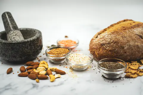 Lasikulhoissa pellavansiemeniä, punaisia linssejä, kaurahiutaleita ja chiansiemeniä, vieressä leipä ja pähkinöitä marmorilla