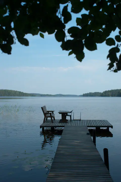 Puulaituri, jonka päässä on penkki, pöytä ja tuoli, tyynellä metsän reunustamalla järvellä kirkkaana kesäpäivänä